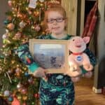 A girl with red hair and glasses in pjs in front of a Christmas tree with her Christmas present, which is a sponsorship certificate and a toy pig for her sponsored pig named Wendell
