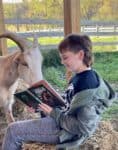 a smiling kid sitting on a bail of straw, holding a book and reading to a goat with horns who is looking into the book.